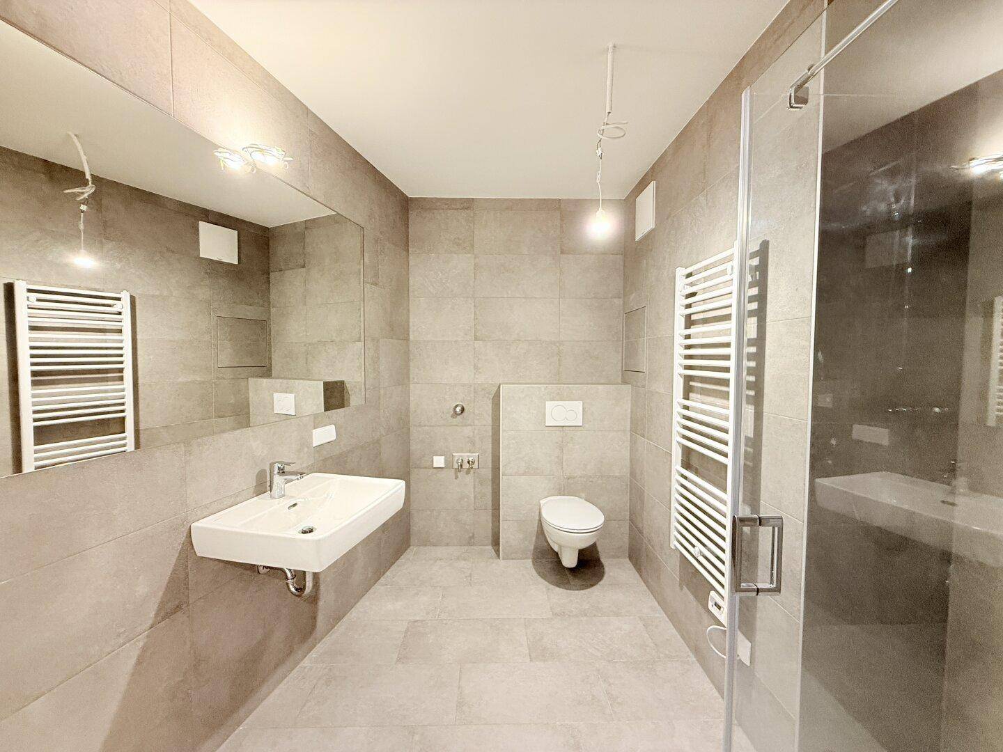 Modernes Badezimmer mit grauen Fliesen, Dusche, WC und beheiztem Handtuchhalter.