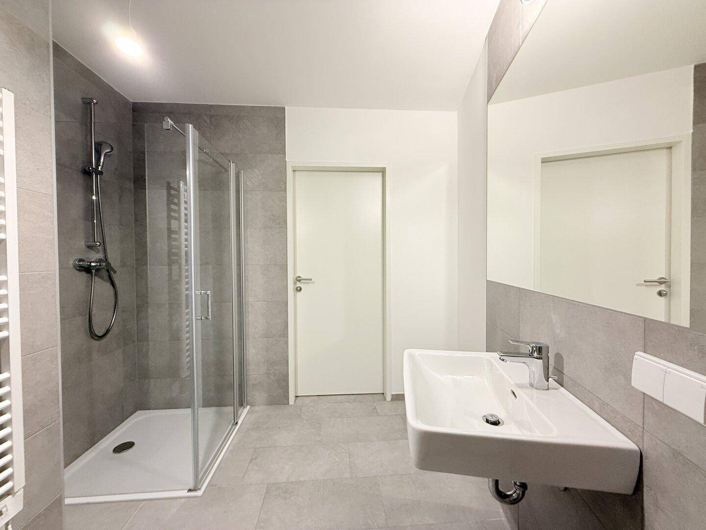 Modernes Badezimmer mit grauen Fliesen, Duschkabine und großem Spiegel über dem Waschtisch.