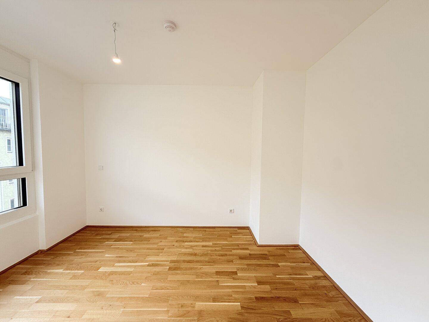 Leeres Zimmer mit Parkettboden, weißen Wänden und viel Tageslicht, ideal als Schlaf- oder Arbeitszimmer.