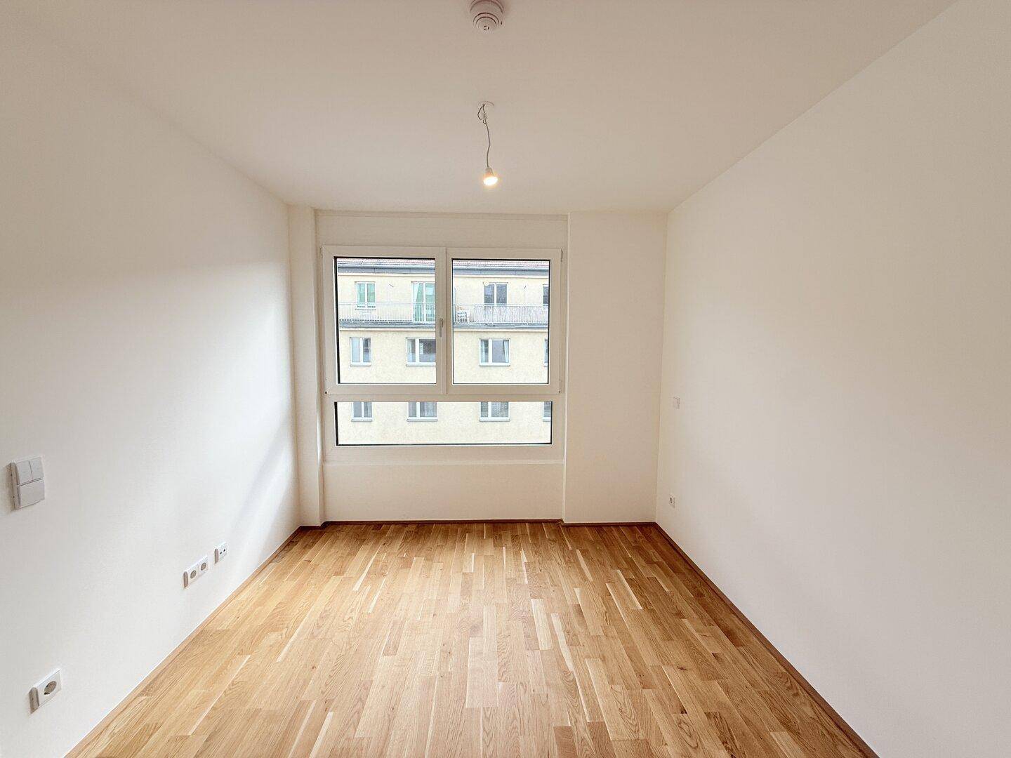 Helles Schlafzimmer mit Parkettboden, großem Fenster und Blick auf die Nachbarschaft.