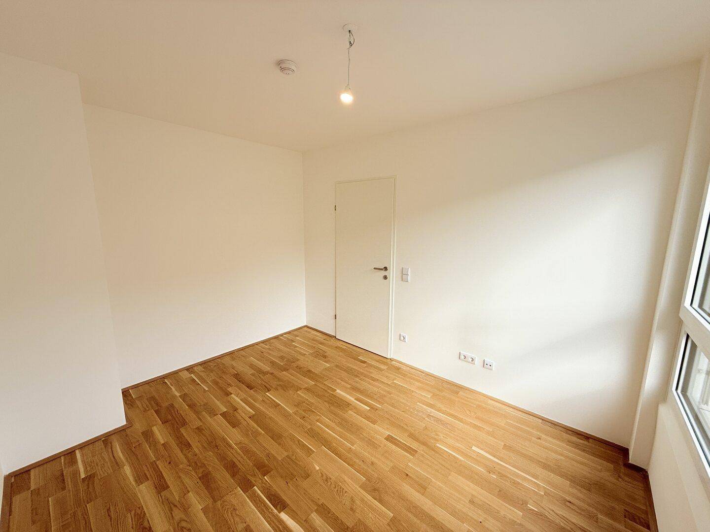 Unmöbliertes Zimmer mit Parkettboden, weißer Tür und Steckdosen an der Wand, bereit zur Nutzung.