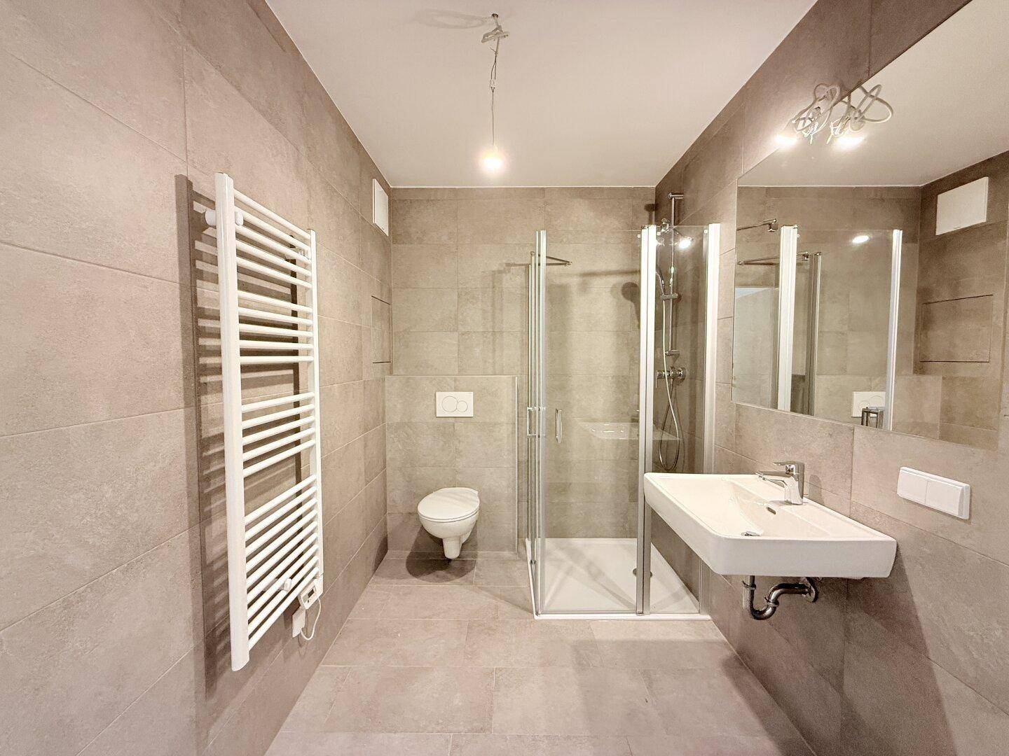 Modernes Badezimmer mit grauen Fliesen, Dusche, Wand-WC und beheiztem Handtuchhalter.