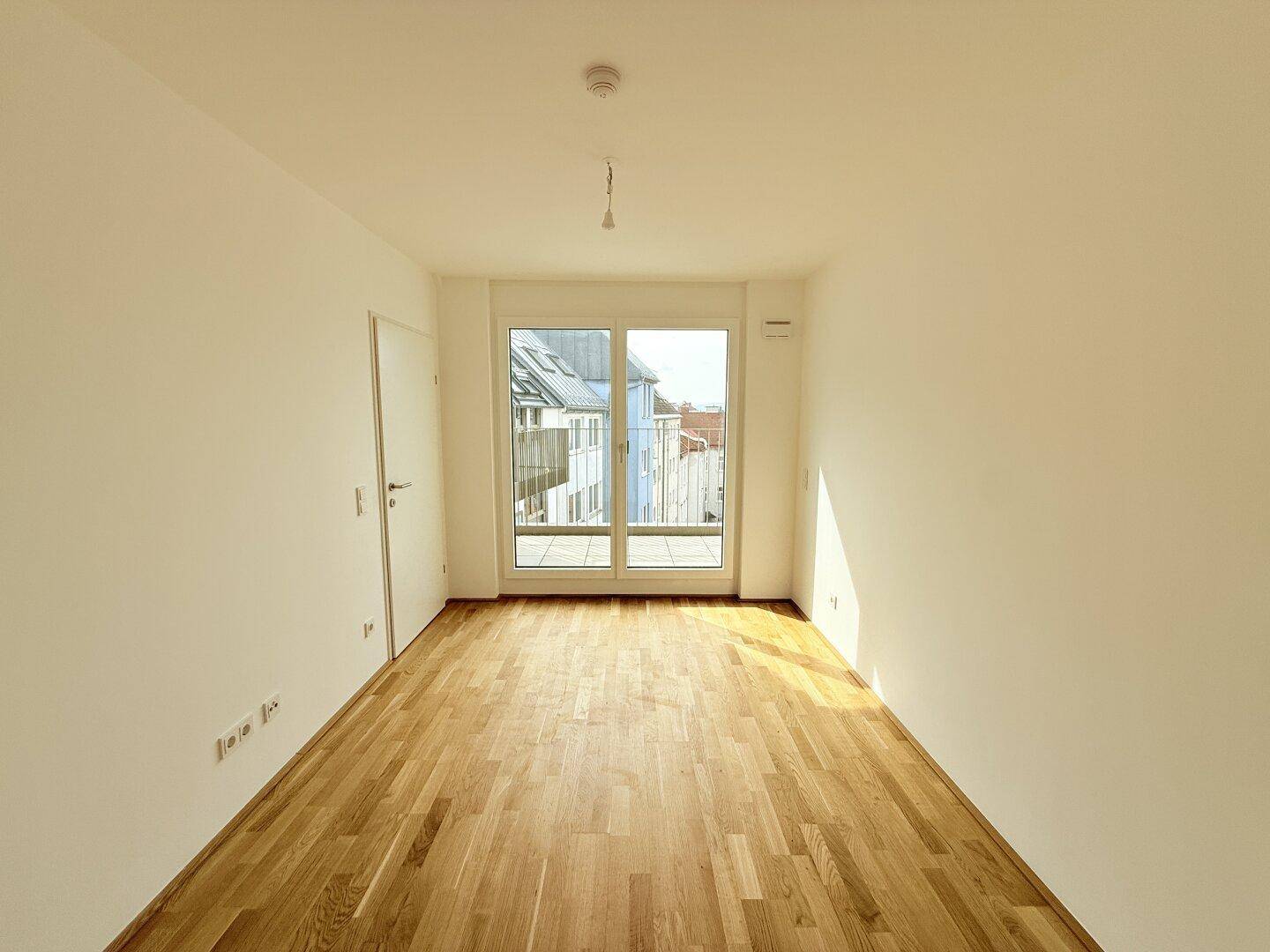 Heller Wohnbereich mit Parkettboden und breiter Fensterfront, die den Zugang zum Balkon ermöglicht.