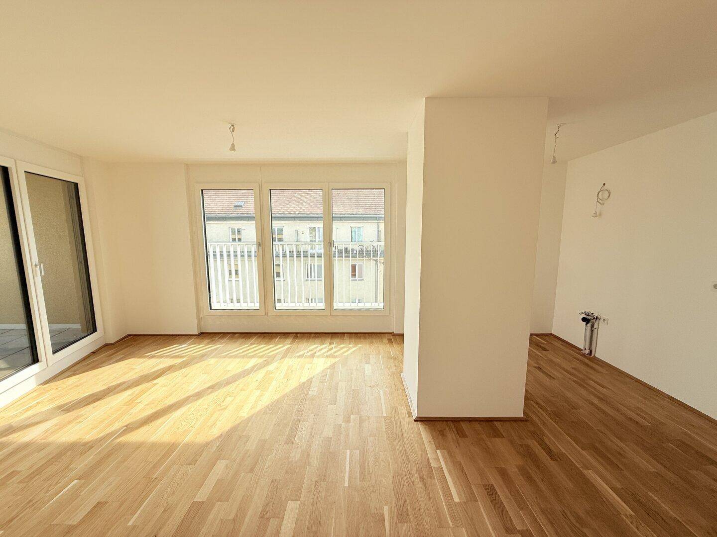 Geräumiges Wohnzimmer mit hellem Parkettboden und großen Fenstern, die viel Tageslicht hereinlassen.