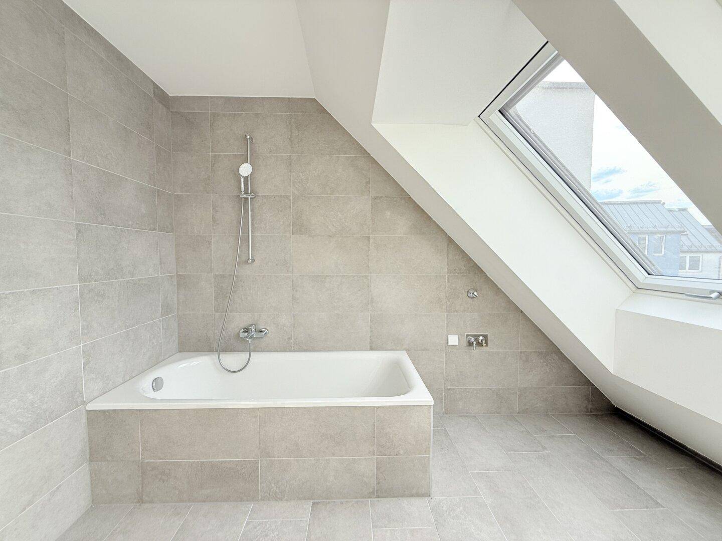 Modernes Badezimmer mit Badewanne, Duscharmatur und grauen Fliesen im Dachgeschoss.