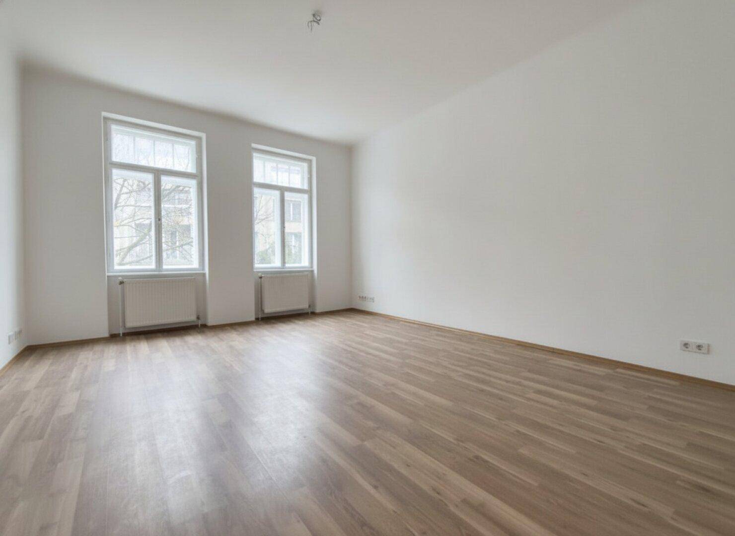 Geräumiges, helles Zimmer mit zwei großen Fenstern und neuwertigem Laminatboden, ideal für Wohnzwecke.