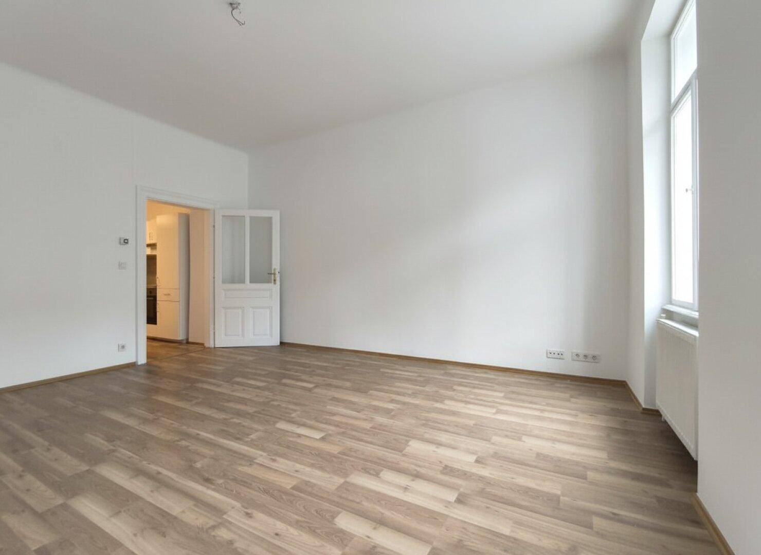 Heller Wohnraum mit Laminatboden und einer Tür, die zu einem weiteren Zimmer führt, mit viel Tageslicht.