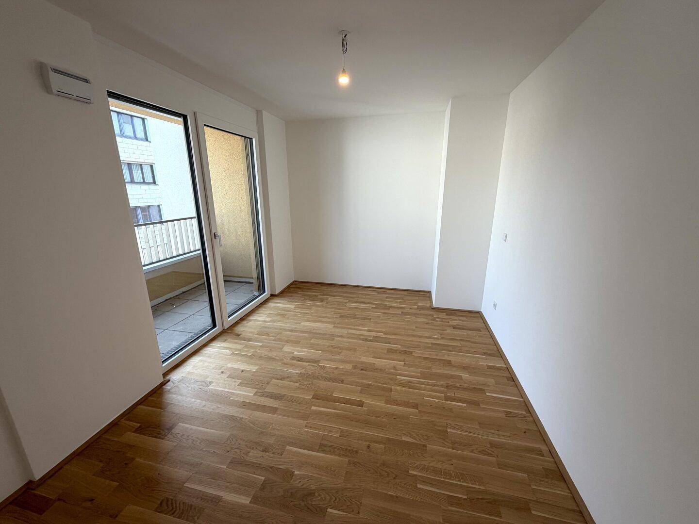 Lichtdurchfluteter Raum mit Parkettboden und Zugang zu einem Balkon.
