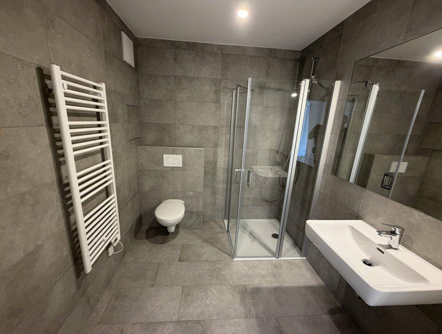 Modernes Badezimmer mit grauen Fliesen, Duschkabine und beheiztem Handtuchhalter.