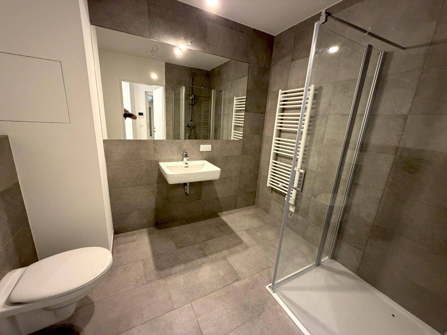 Modernes Badezimmer mit grauen Fliesen, Dusche und beheiztem Handtuchhalter.