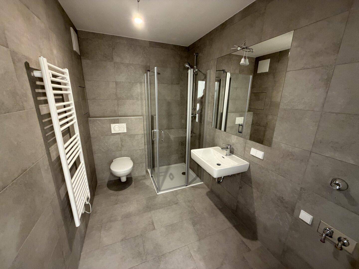 Modernes Badezimmer mit grauen Fliesen, Dusche, WC und beheiztem Handtuchhalter.