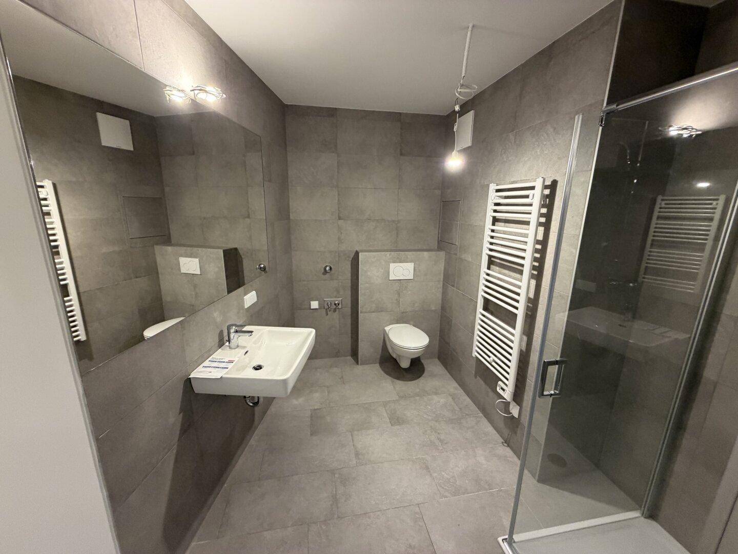 Modernes Badezimmer mit grauen Fliesen, wandhängendem WC und einer geräumigen Duschkabine.