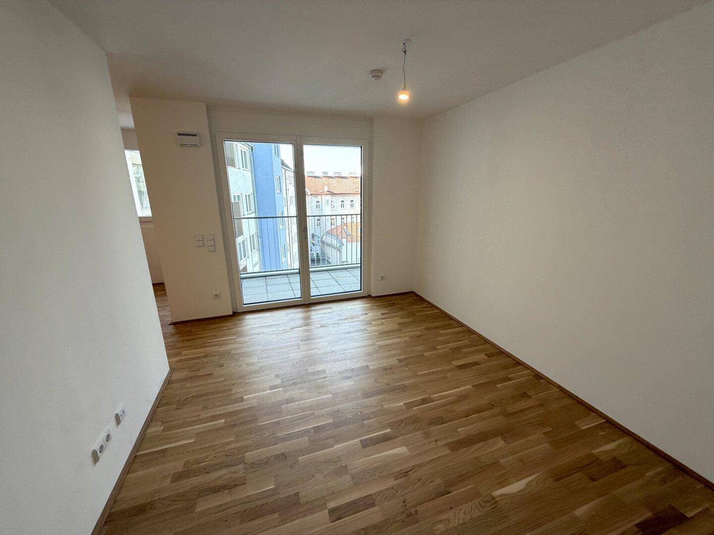 Lichtdurchfluteter Raum mit Holzparkett und großem Fenster, das Zugang zum Balkon bietet.