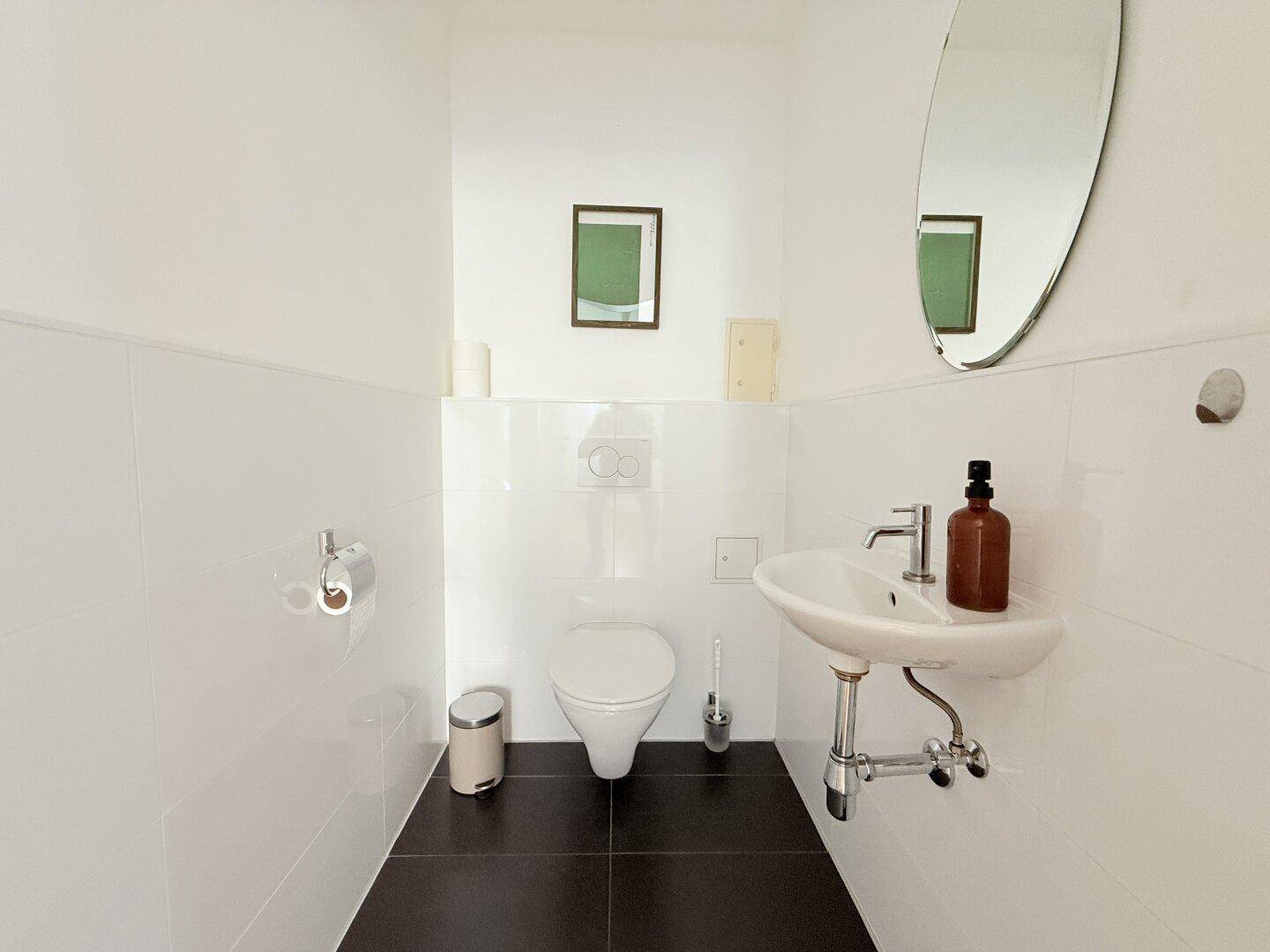 Modernes Gäste-WC mit weißen Wandfliesen, dunklem Boden und ovalem Spiegel.