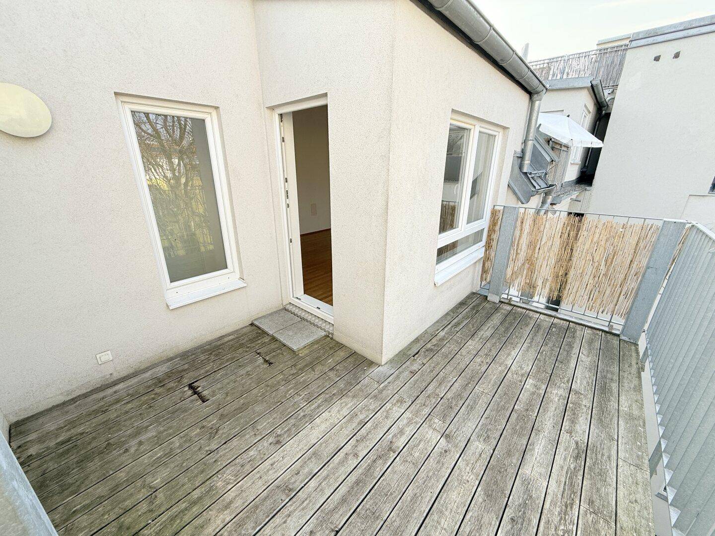 Geräumiger Balkon mit Holzboden und Zugang zum Innenbereich, umgeben von hellen Fassaden.