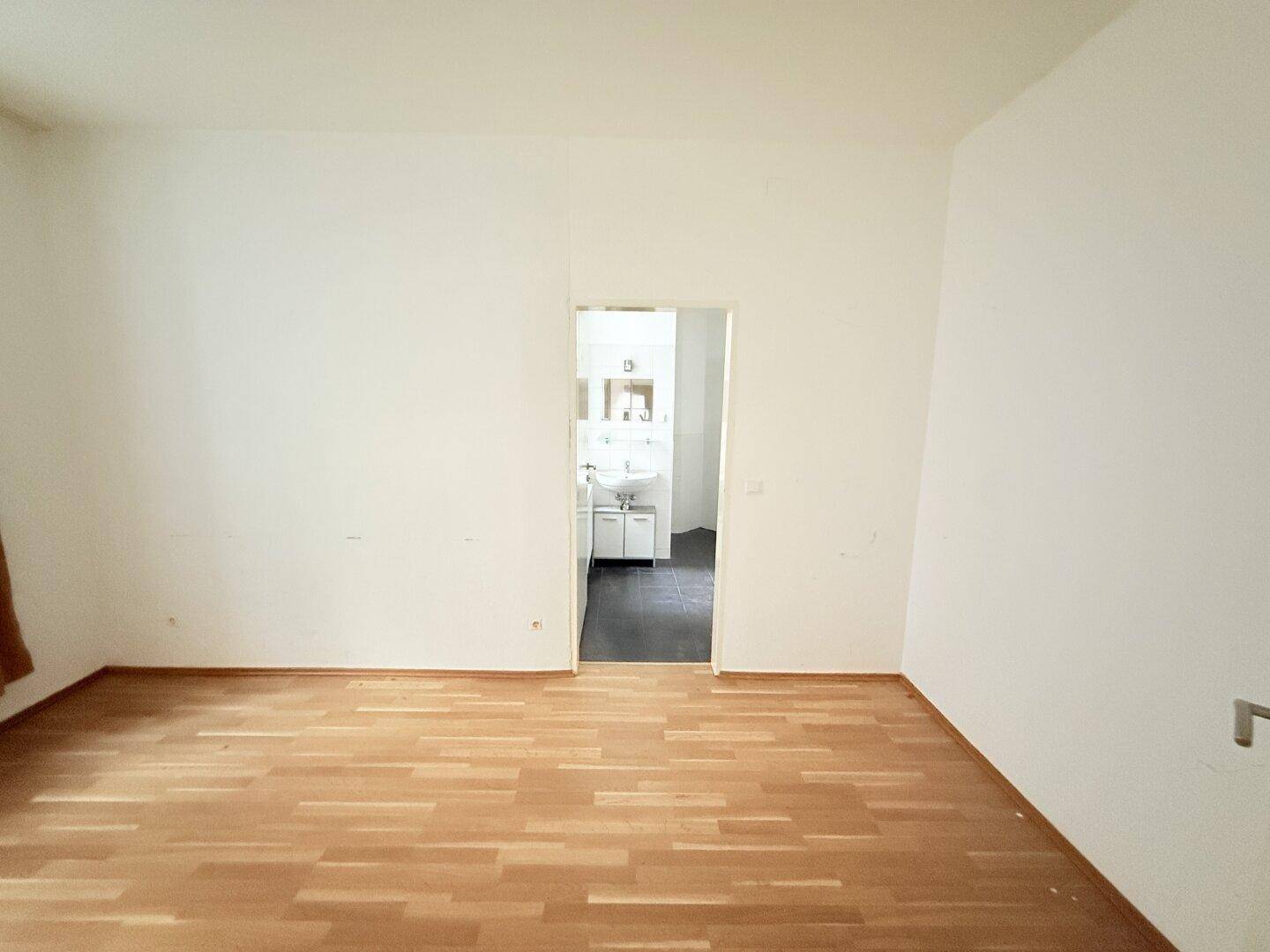 Leerer Raum mit Holzparkettboden und einem Blick durch die Tür in ein Badezimmer.