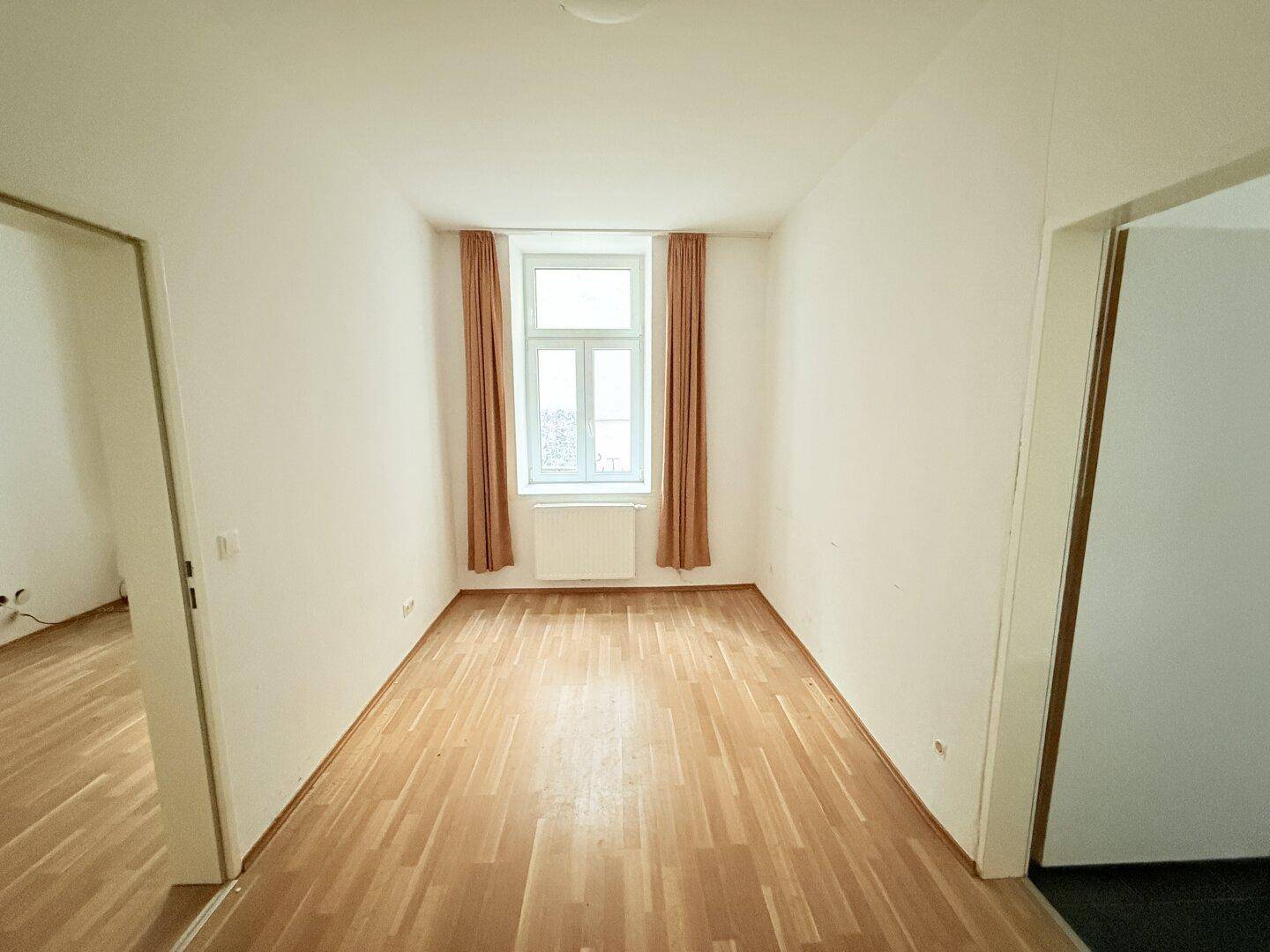 Heller, leerer Raum mit Holzparkettboden, Fenster und braunen Vorhängen.