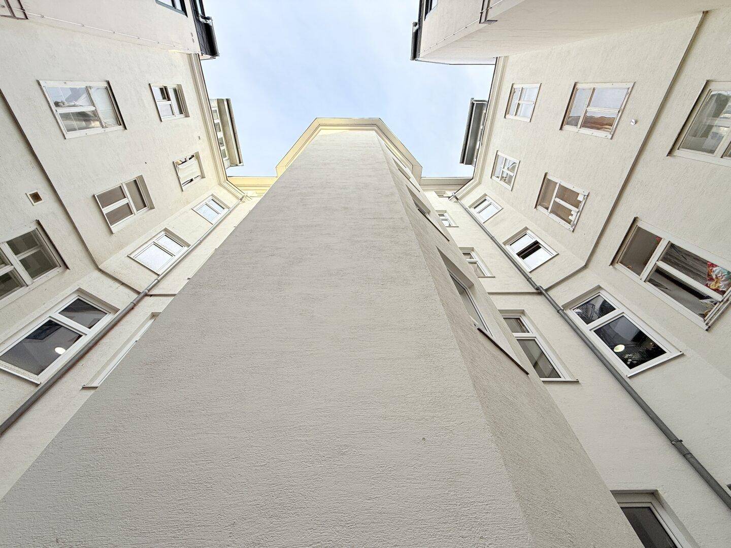 Blick nach oben in einen hellen Innenhof eines Wohngebäudes mit vielen Fenstern.
