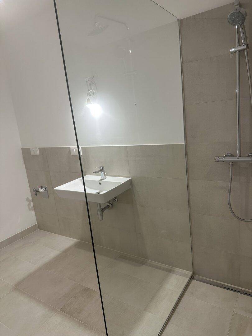 Modernes Badezimmer mit begehbarer Dusche und Waschbecken, hell gefliest und minimalistisch gestaltet.