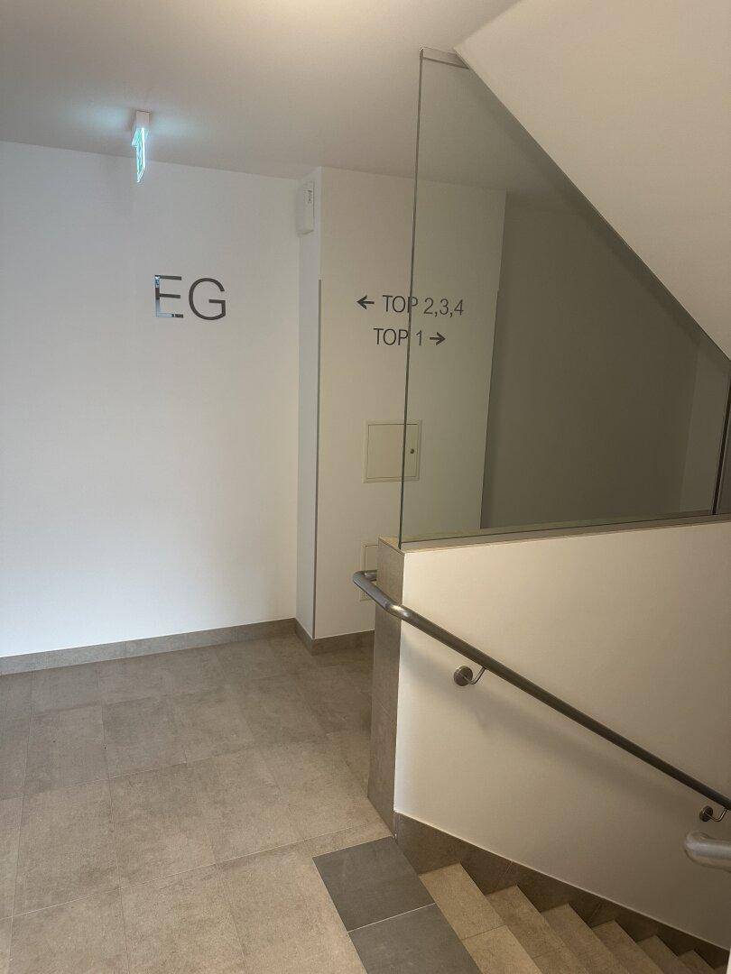 Heller Eingangsbereich mit Treppe, Glasgeländer und Hinweisschildern zu den verschiedenen Wohneinheiten.