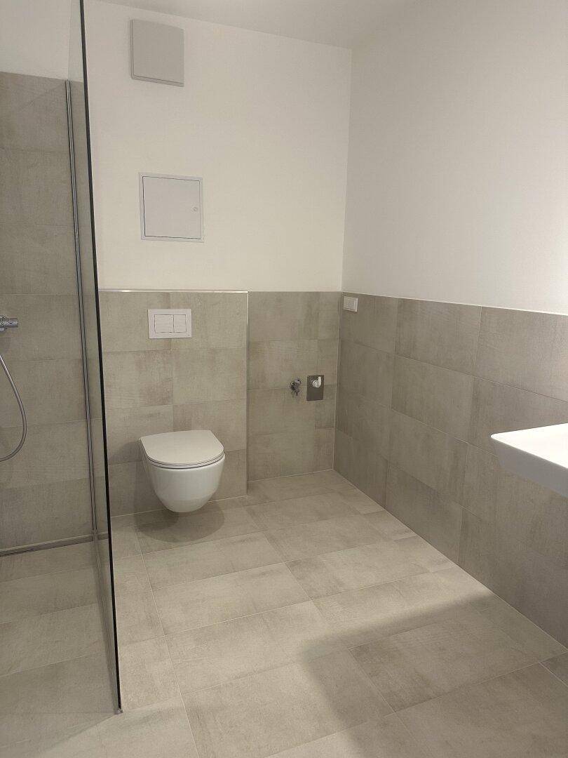 Modernes Badezimmer mit grauen Fliesen, wandhängendem WC und einer geräumigen begehbaren Dusche.
