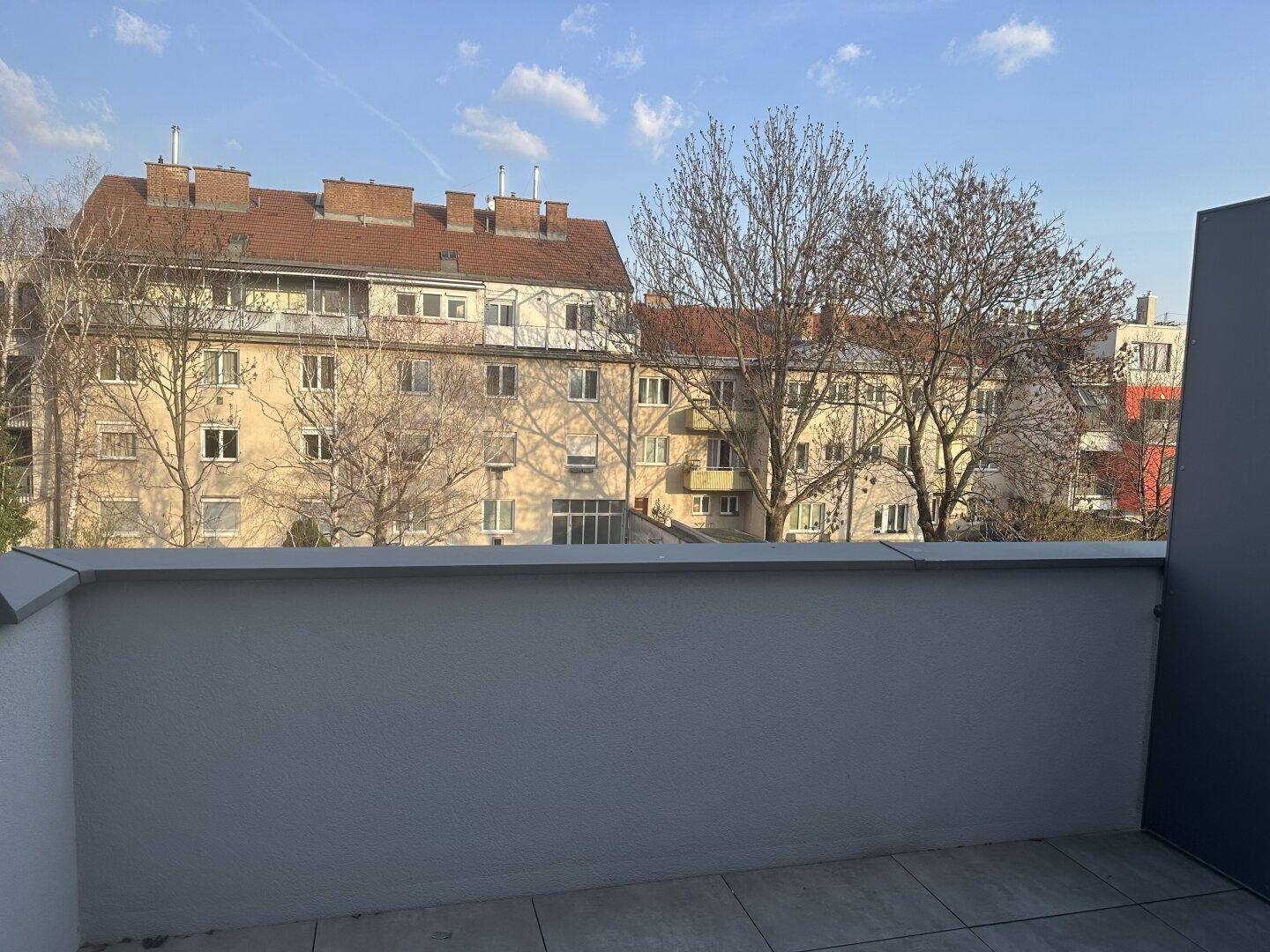 Ausblick von einem Balkon auf umliegende Gebäude und Bäume unter einem klaren blauen Himmel.