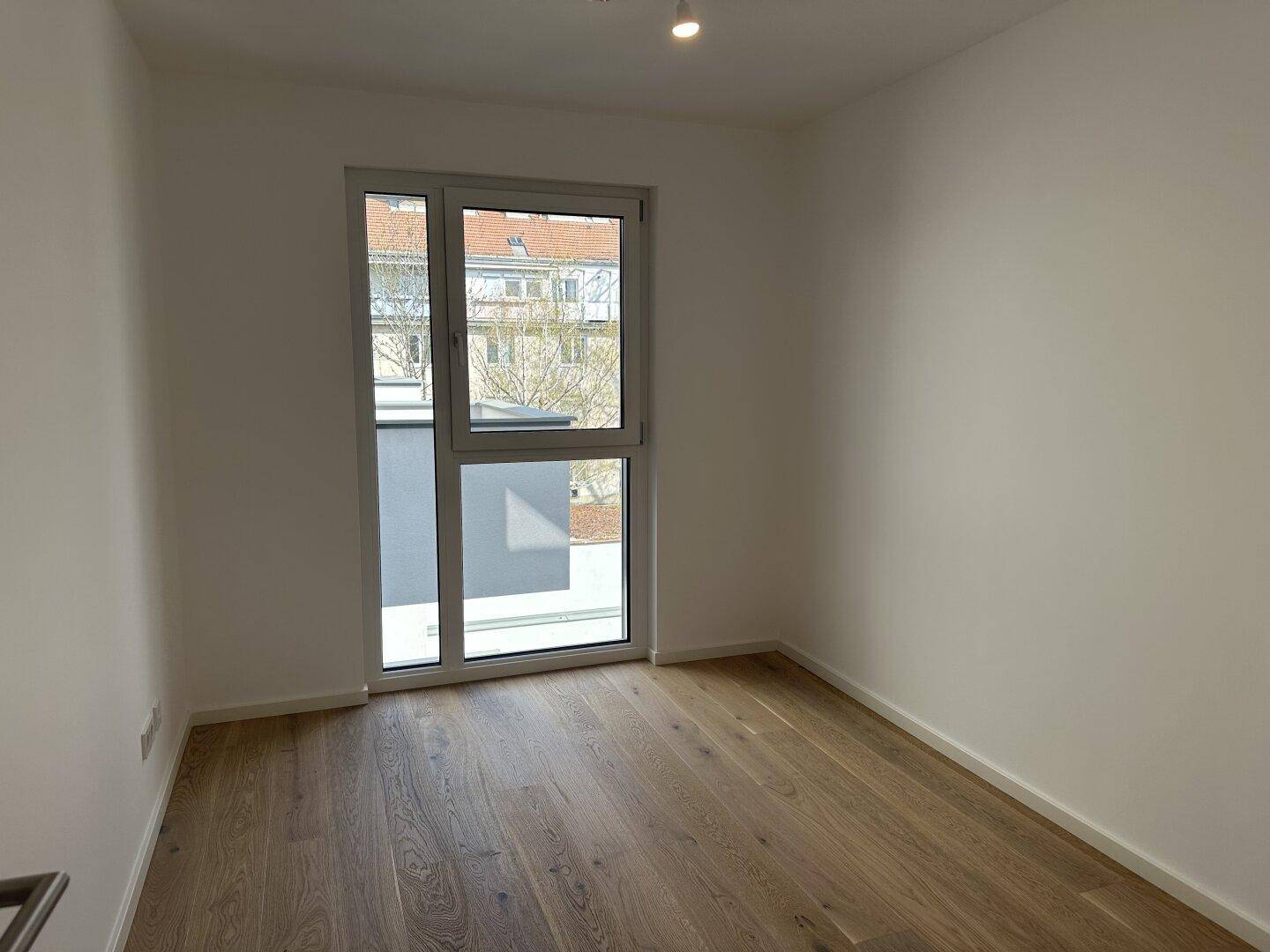 Helles Zimmer mit Holzboden und großem Fenster, das viel Tageslicht hereinlässt, ideal als Schlafzimmer.