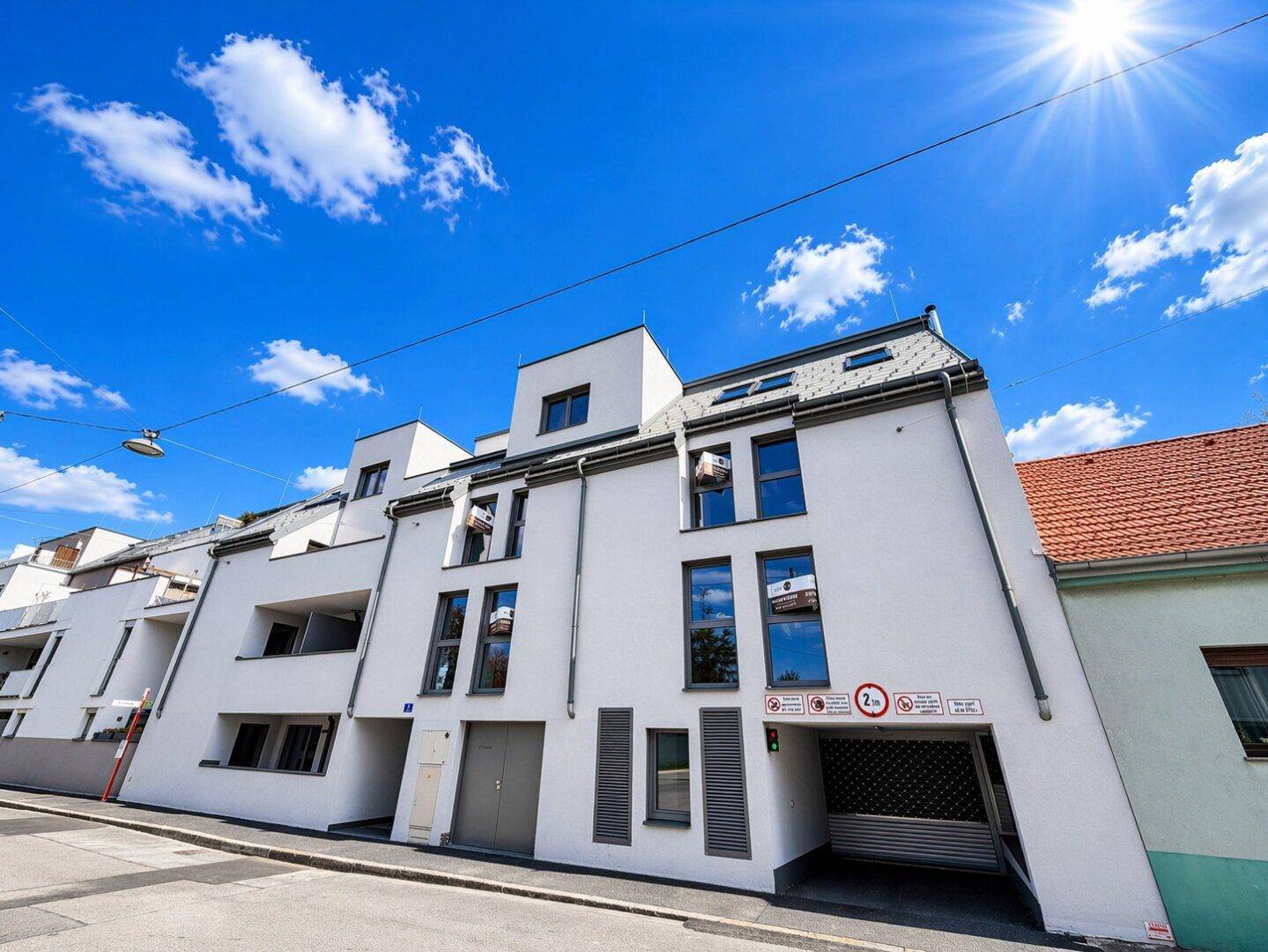 Neubau mit weißer Fassade, großen Fenstern und Tiefgaragenzufahrt unter strahlend blauem Himmel.