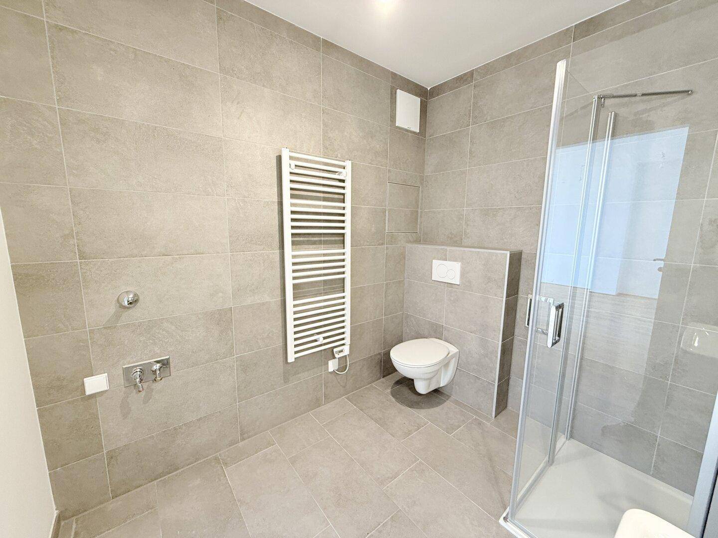 Zeitgemäßes Badezimmer mit Dusche, WC und Handtuchheizkörper in neutralen Grautönen.