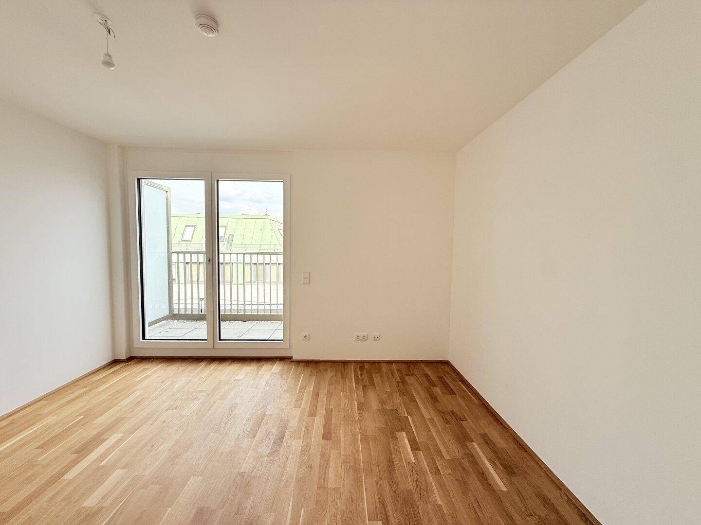 Leeres, helles Zimmer mit Holzparkettboden und großem Fenster, das Zugang zu einem Balkon bietet.