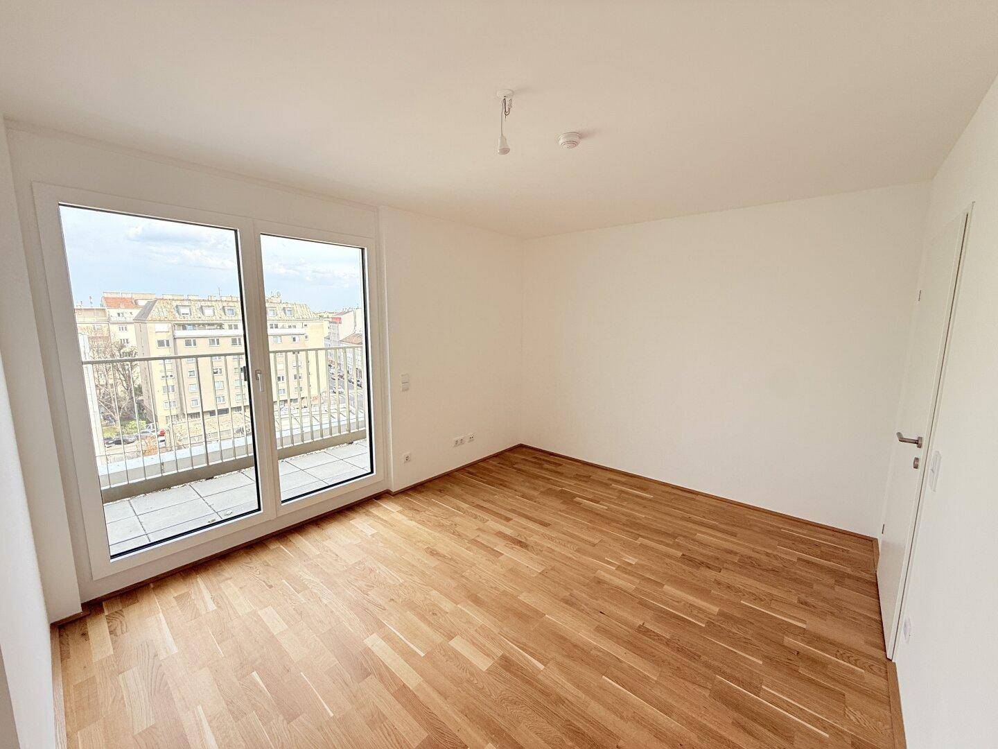Helles Zimmer mit Holzparkettboden und großem Fenster, das den Zugang zu einem Außenbereich ermöglicht.