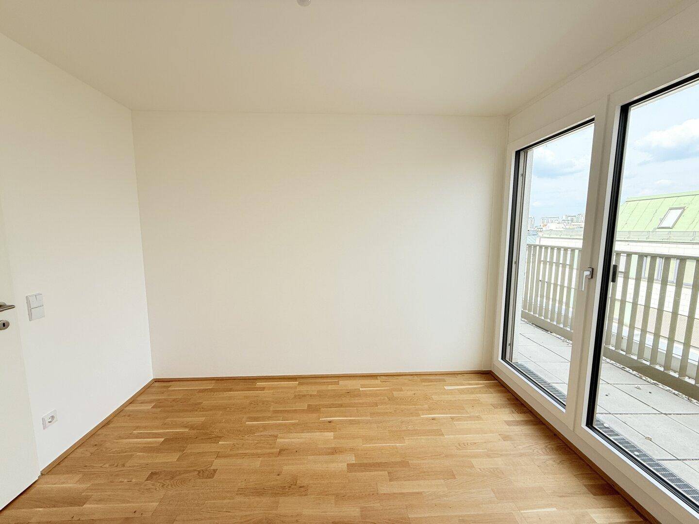 Helles Zimmer mit Holzparkettboden und bodentiefen Fenstern, die den Blick auf einen Balkon freigeben.