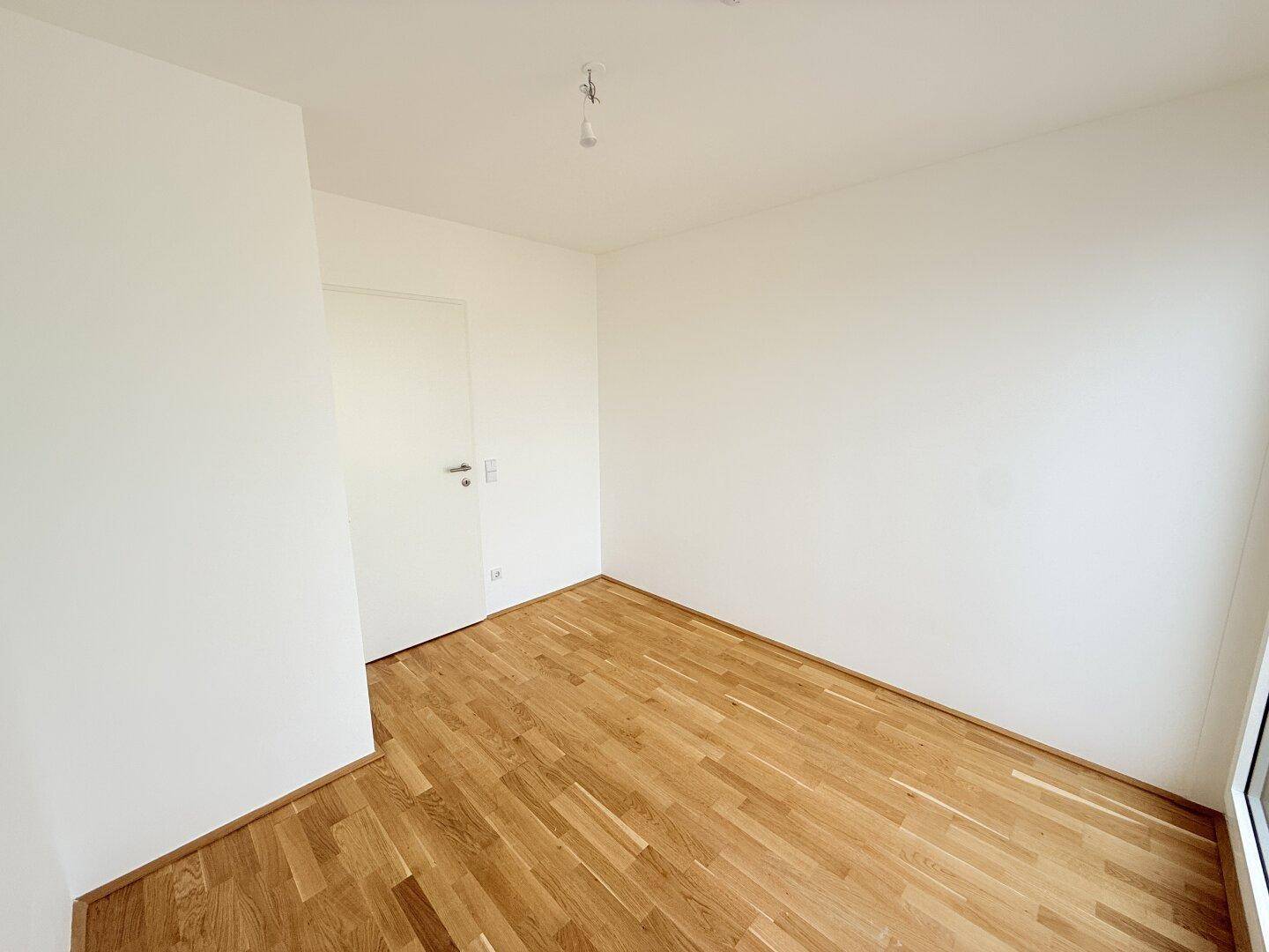 Leeres Zimmer mit Holzparkettboden und weißen Wänden, das eine Tür und einen hellen Fensterbereich zeigt.