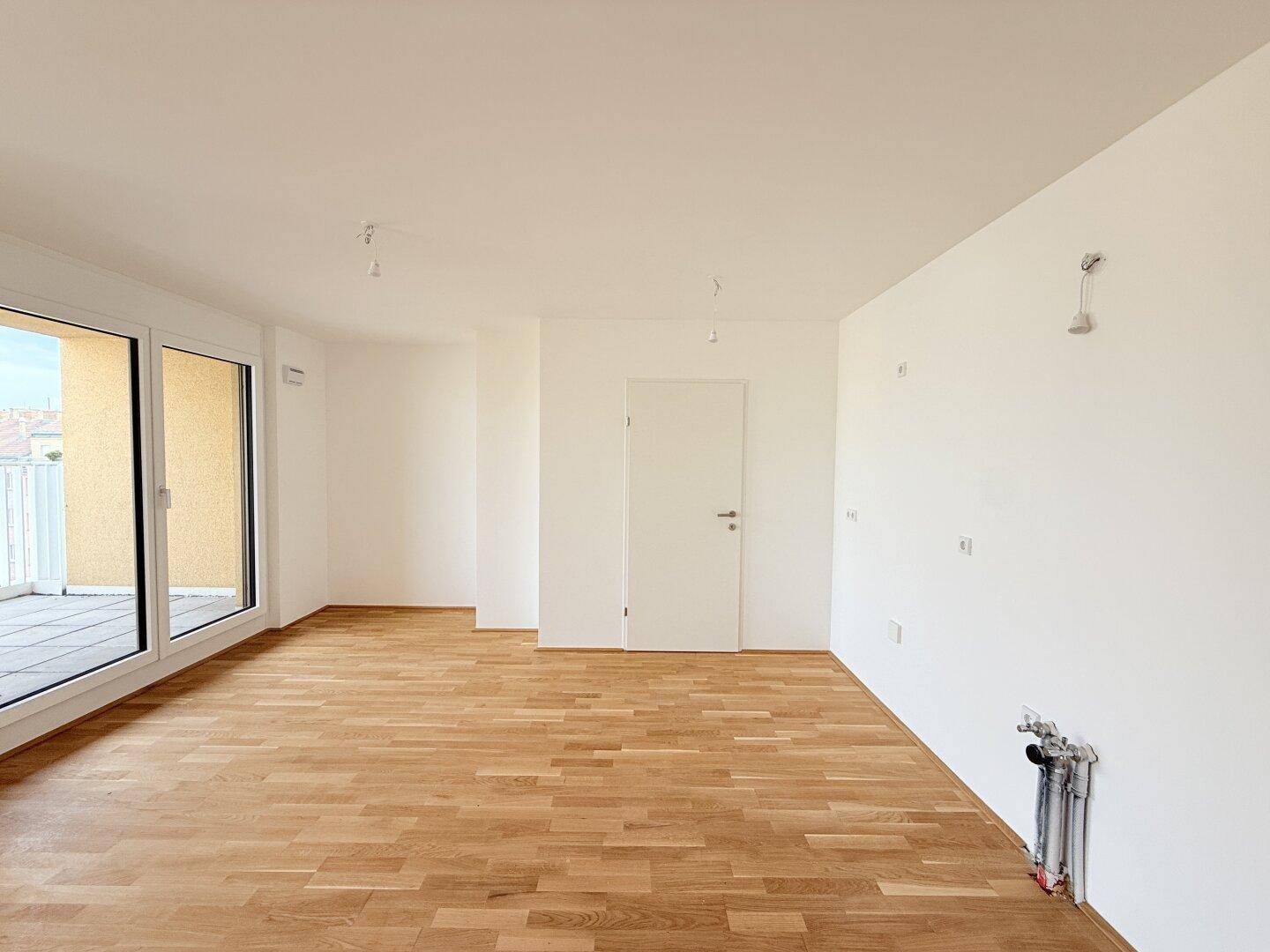 Leerer Raum, vorgesehen als Küche, mit Holzparkettboden und sichtbaren Anschlüssen für Küchengeräte.
