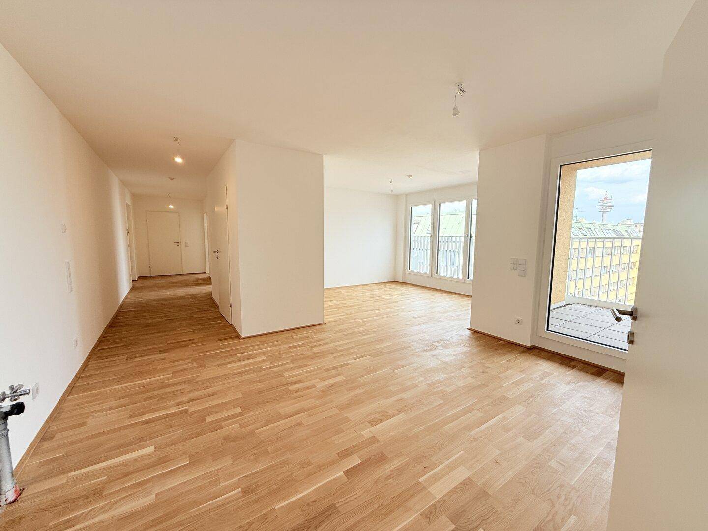 Offener Wohnbereich mit Holzparkettboden und großen Fenstern, der in einen Flur übergeht.