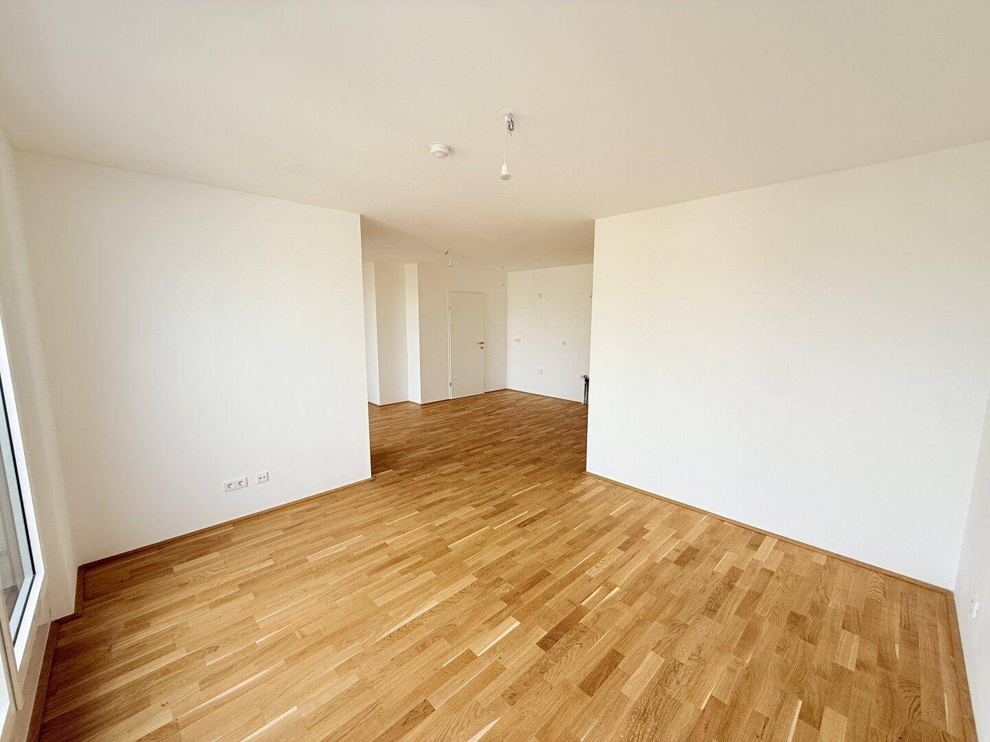 Geräumiger Wohnbereich mit Holzparkettboden und weißen Wänden, der offen zu einem weiteren Raum ist.