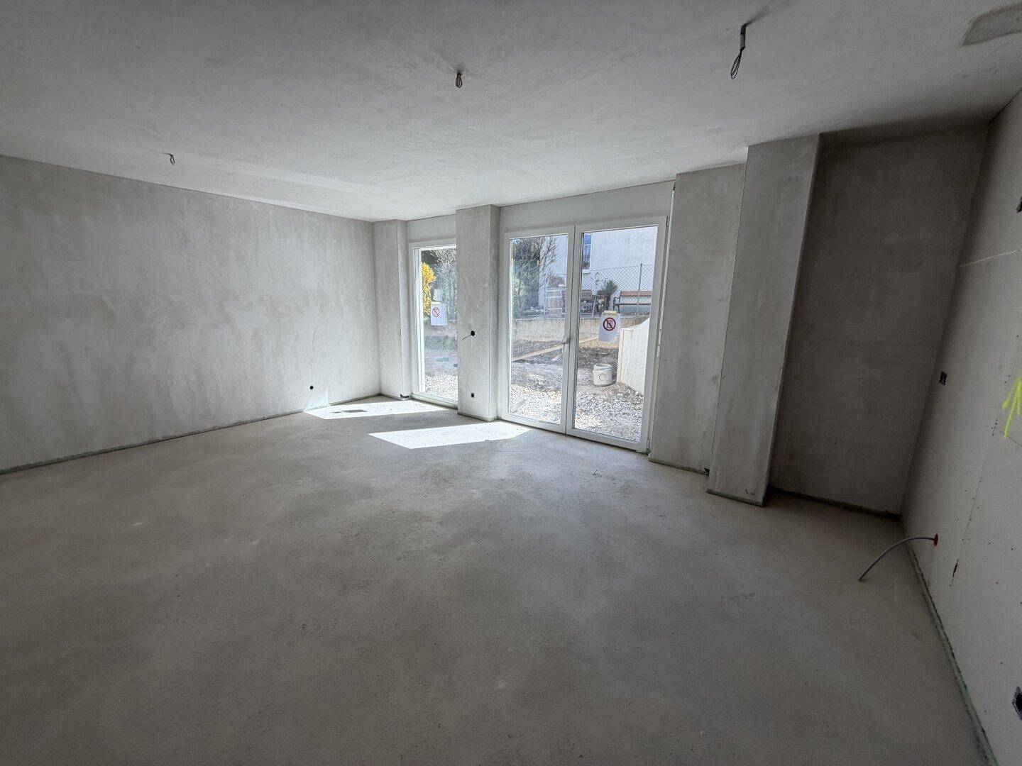 Heller, unmöblierter Raum im Rohbau mit zwei großen Fensterfronten und Betonboden.