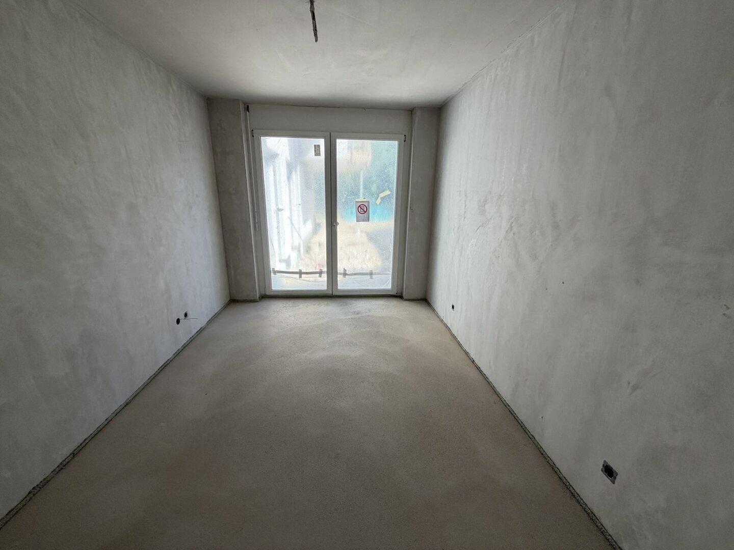 Unfertiger Innenraum mit Betonboden und großen Glastüren, bereit für den weiteren Ausbau.