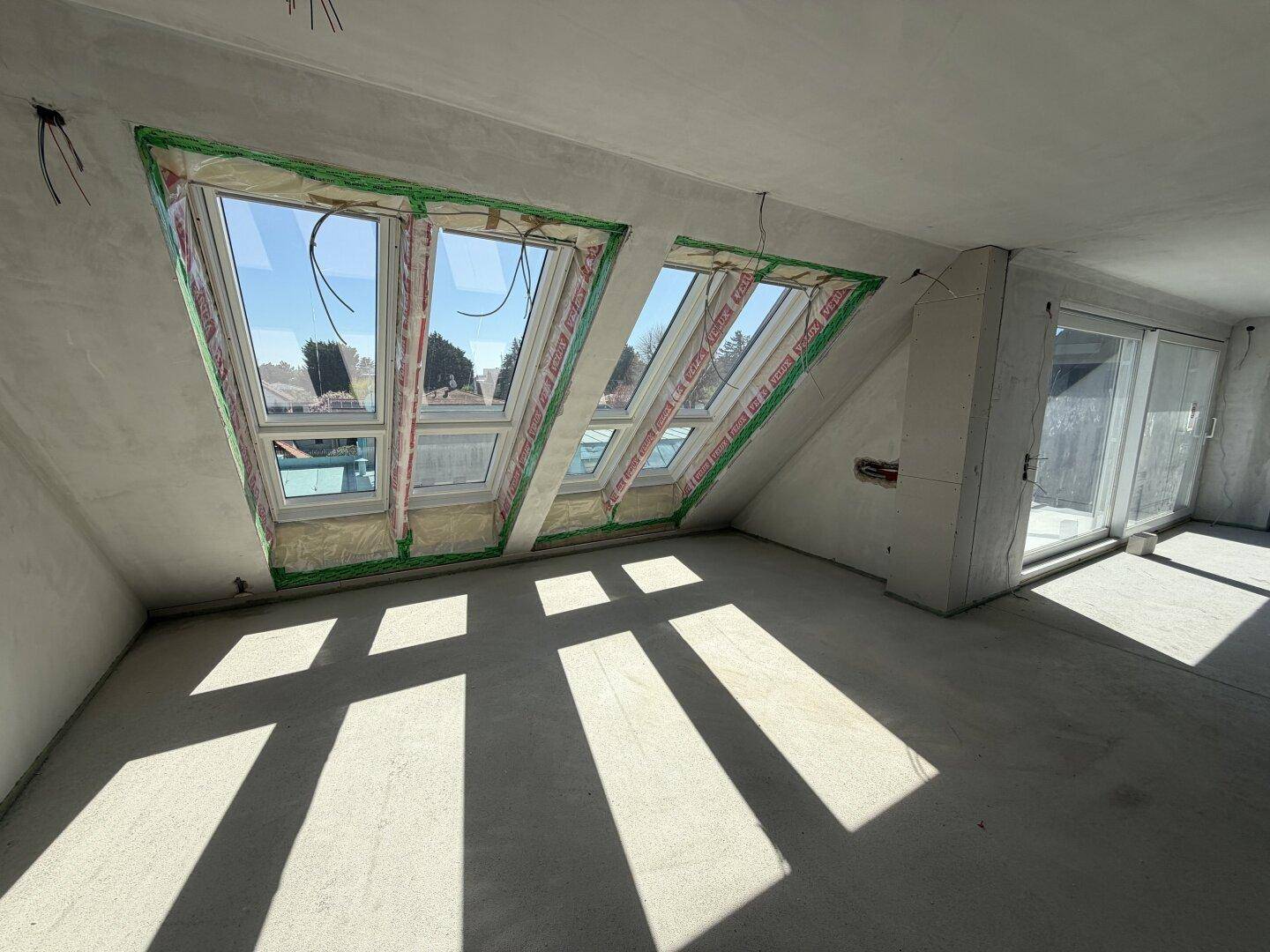 Heller Rohbau-Raum mit großen Dachfenstern und Zugang zu einem potenziellen Außenbereich.