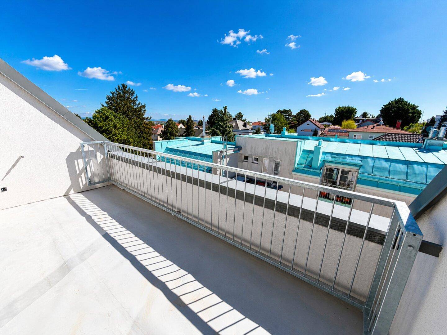 Heller Balkon mit Blick auf die Dächer der Nachbarschaft und grüne Bäume unter blauem Himmel.