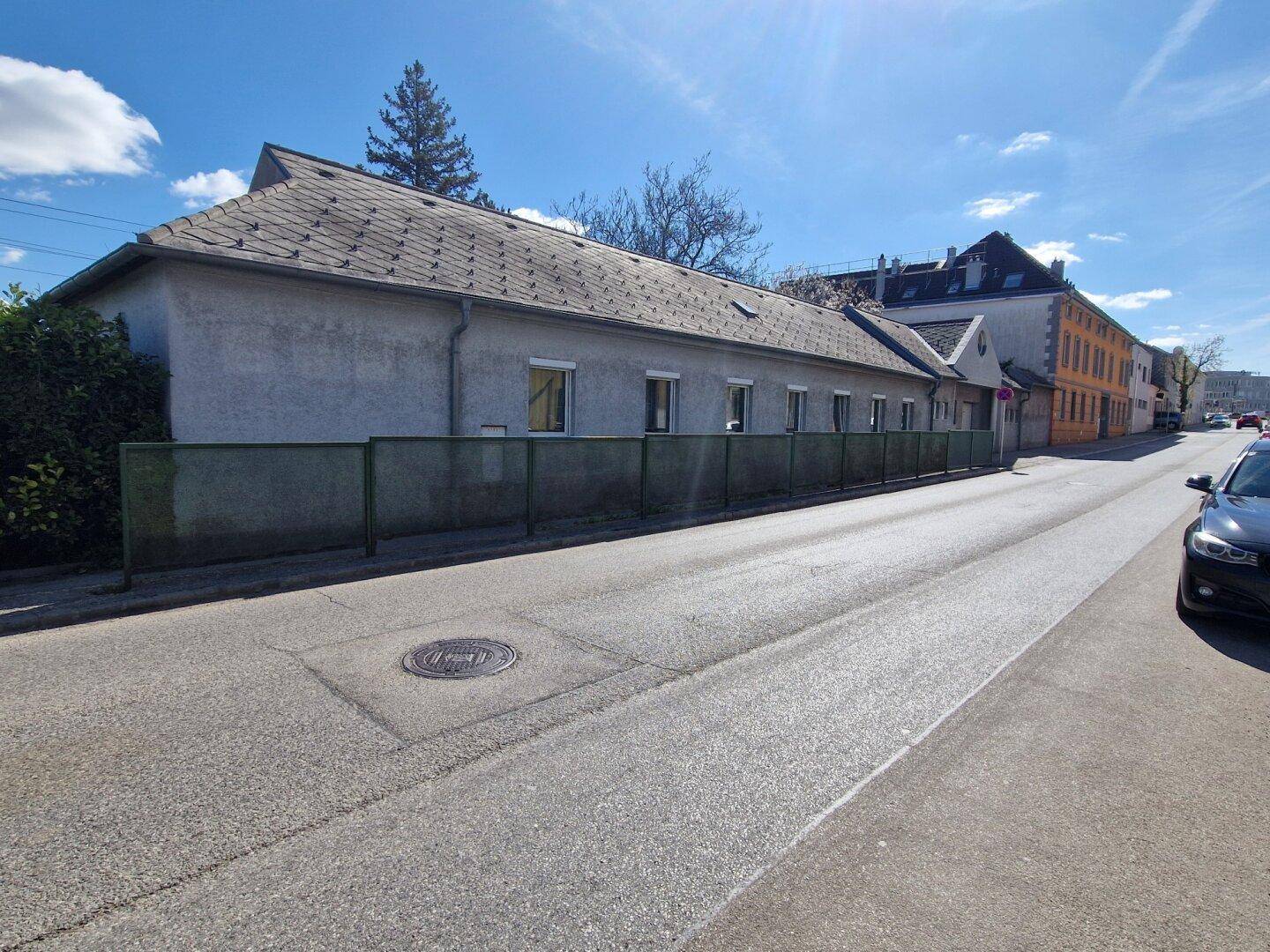 Längliche Fassade eines Gebäudes entlang einer ruhigen Straße mit einem grünen Zaun und klarem Himmel.