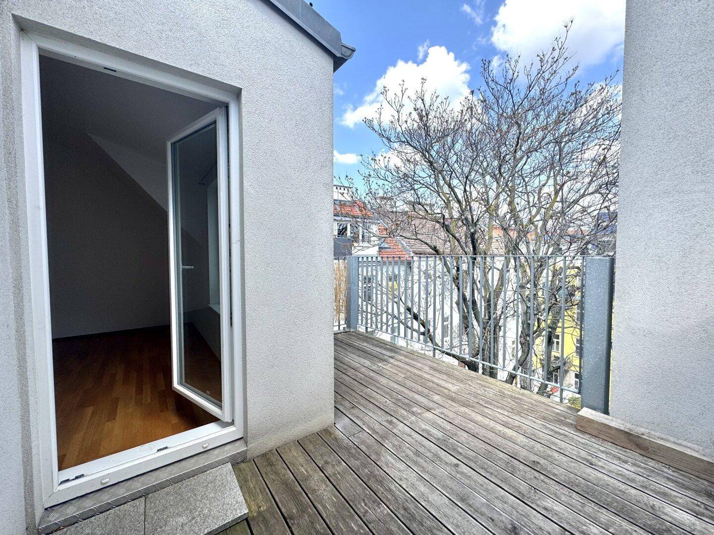 Sonniger Balkon mit Holzboden und Zugang zum Innenbereich, umgeben von Bäumen und Stadtansicht.