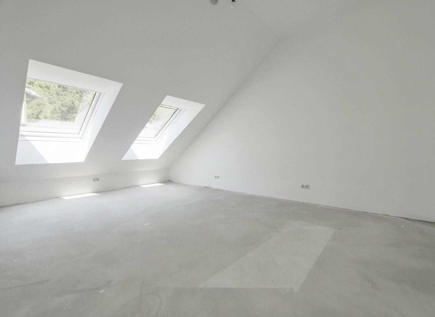 Heller Dachgeschossraum im Rohbau mit Dachfenstern und schrägen Wänden.