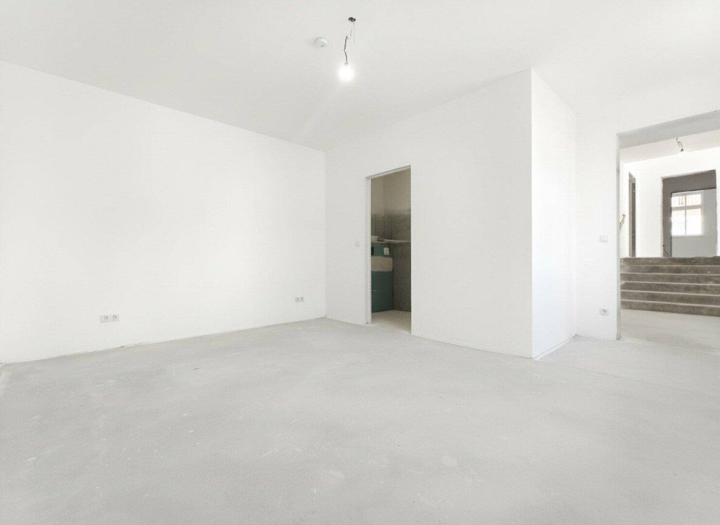 Heller Rohbau-Raum mit Betonboden und Blick auf eine Treppe.