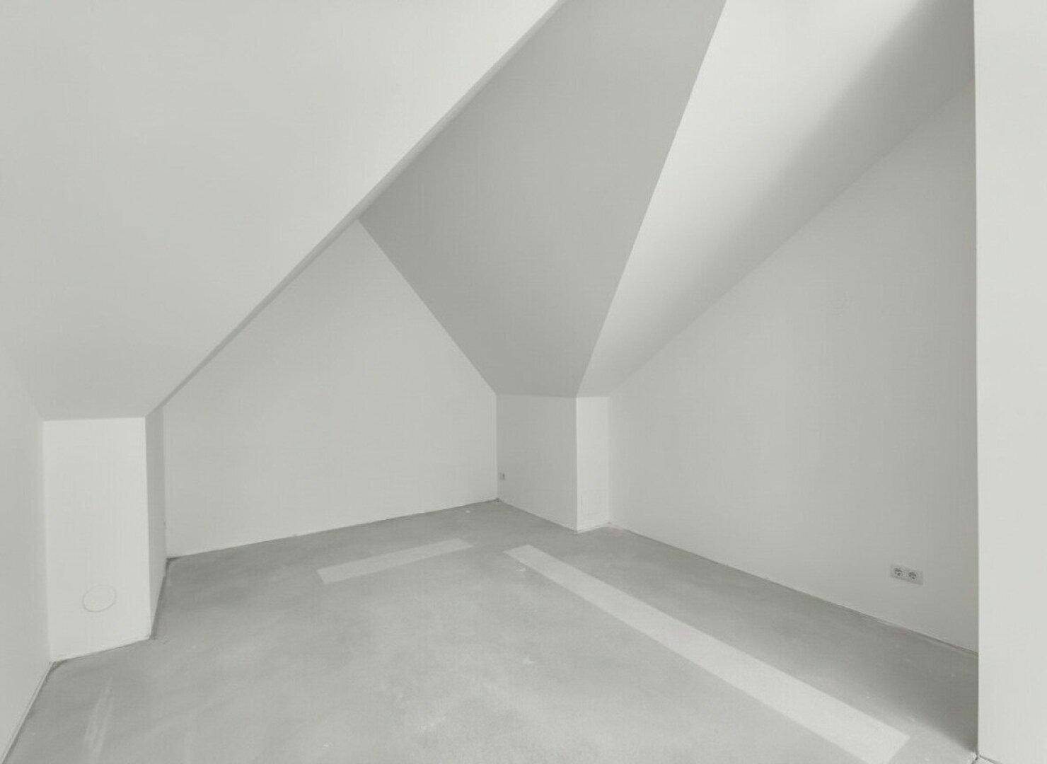 Geräumiger Dachgeschossraum im Rohbau mit schrägen Wänden und Betonboden.