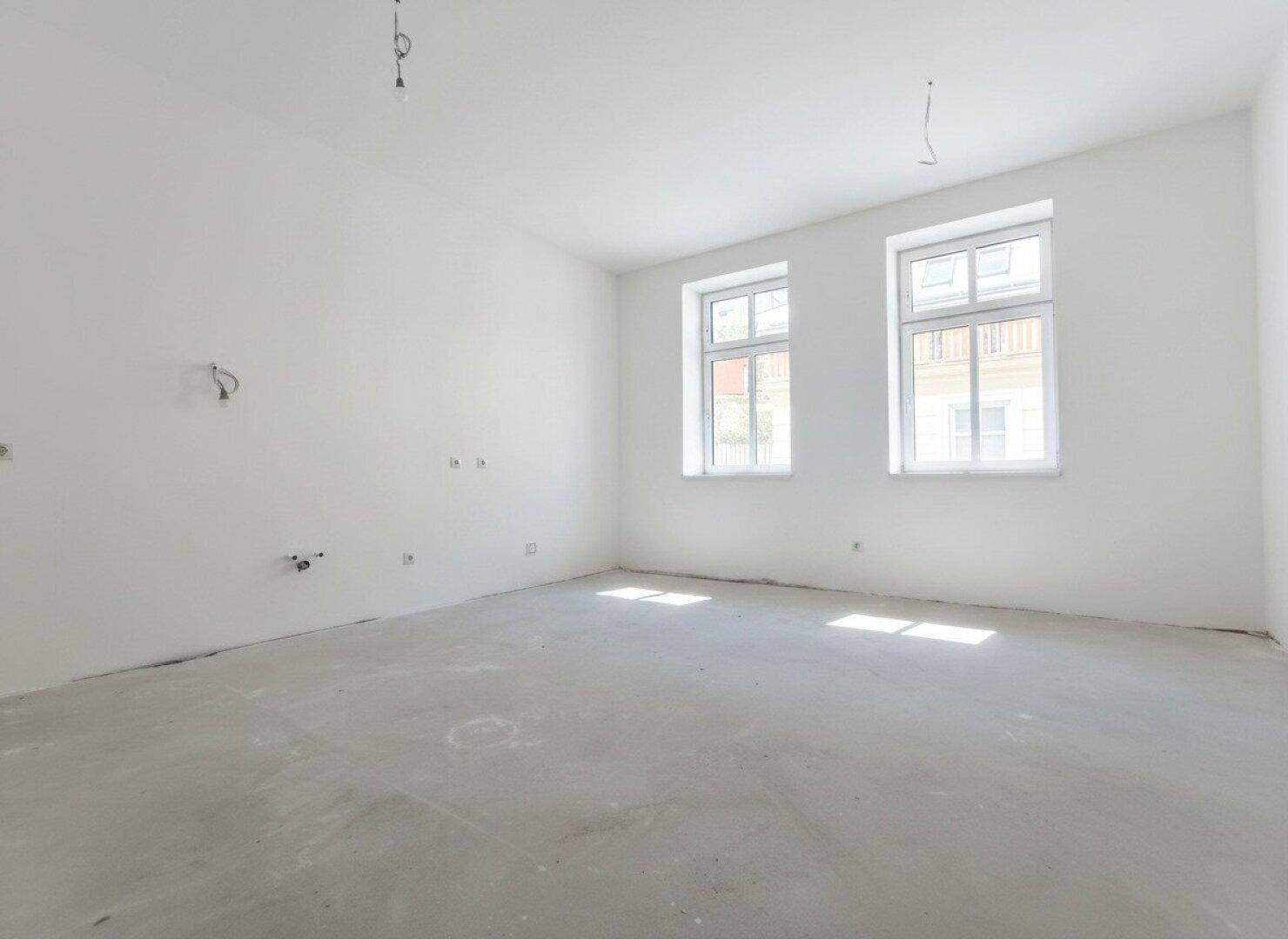 Leerer Raum im Rohbau mit zwei Fenstern und Betonboden.