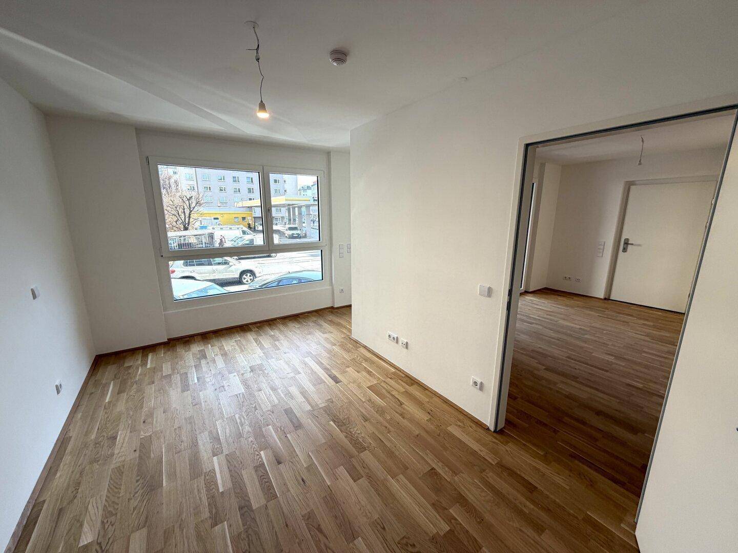 Heller Raum mit Holzboden, weißen Wänden und großem Fenster mit Blick auf die Straße.
