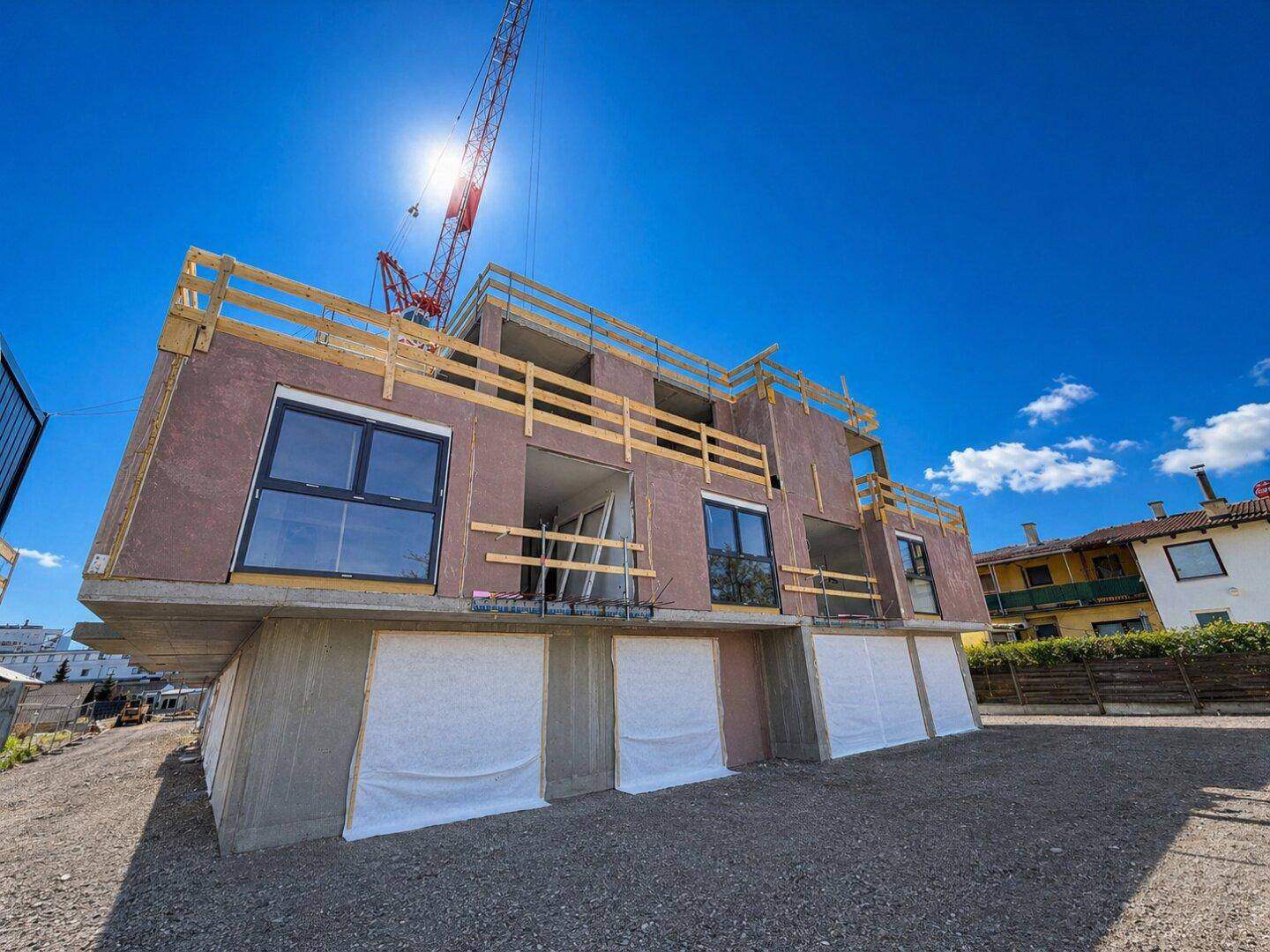 Neubau im Bau mit großen Fensteröffnungen und einem Baukran unter strahlendem Sonnenschein.