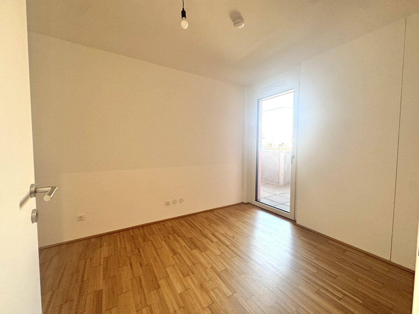 Geräumiges Schlafzimmer mit Holzboden und viel Tageslicht durch die Balkontür.
