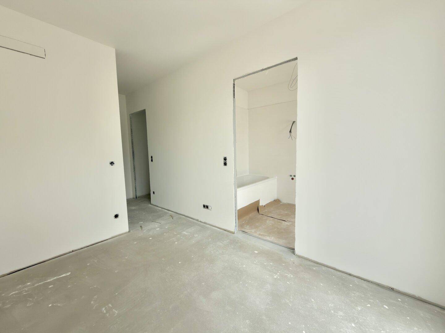 Unmöblierter Raum im Rohbauzustand mit weiß gestrichenen Wänden und Blick in ein Badezimmer.